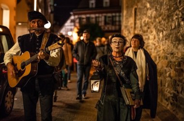 Ein gewandeter Mann mit Gitarre sowie eine gewandete und weiß geschminkte Frau mit Laterne in der Hand laufen durch die dunklen Gassen. Im Hintergrund sind weitere Personen zu sehen.