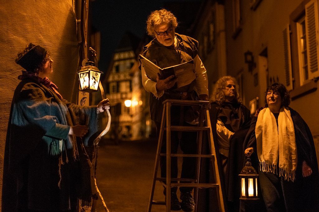 Laternenführung Vier gewandete Personen bei Nacht sind zu sehen. Eine Person im Hintergrund. Die Frauen links und rechts im Vordergrund halten beleuchtete Laternen, der Mann in der Mitte steht auf einer Holzleiter und liest aus einem Buch.