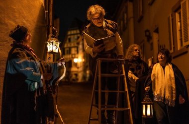 Vier gewandete Personen bei Nacht sind zu sehen. Eine Person im Hintergrund. Die Frauen links und rechts im Vordergrund halten beleuchtete Laternen, der Mann in der Mitte steht auf einer Holzleiter und liest aus einem Buch.