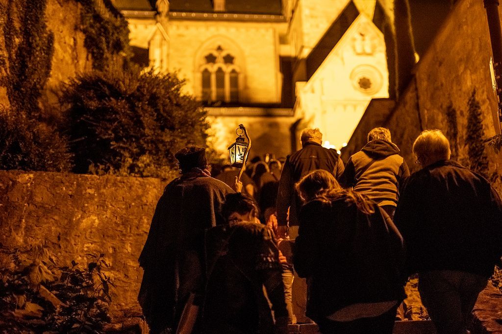Laternenführung Eine Gruppe Personen läuft bei Nacht auf die im Hintergrund beleuchtete Kirche zu. Links hält eine gewandete Gästeführerin eine Laterne an einem Holzstab.