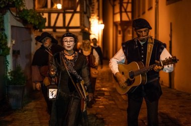 Ein gewandeter Mann mit Gitarre links und eine gewandete und weiß geschminkte Frau mit Laterne laufen bei Nacht durch die Gassen.