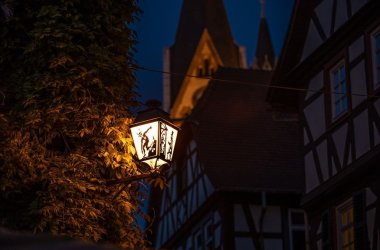 Beleuchtete Laterne mit Scherenschnitt an Häuserwand bei Nacht. Der Scherenschnitt zeigt den Teufel.